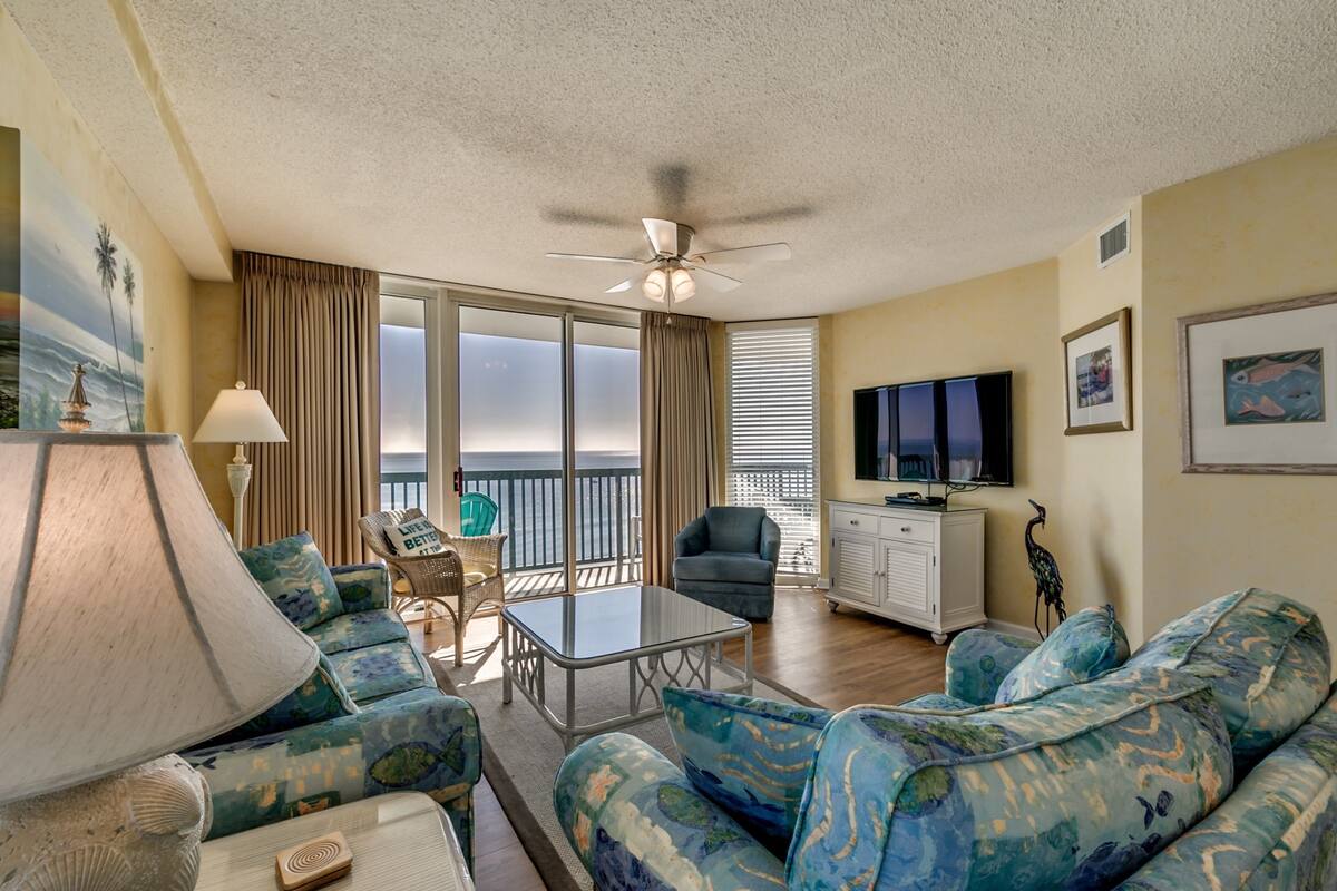 A living room with a sofa, armchair, and a table. There is a ceiling fan, a TV, and a window with curtains.