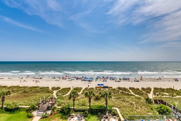 A beach with a wooden walkway leading to a structure, palm trees, and beach umbrellas.