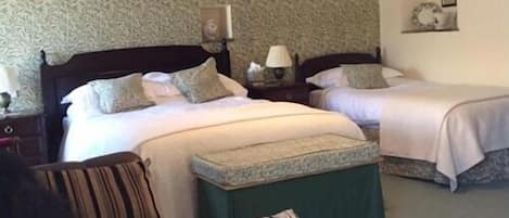 A bedroom with two beds, a nightstand with a lamp, a green ottoman, and a striped chair.
