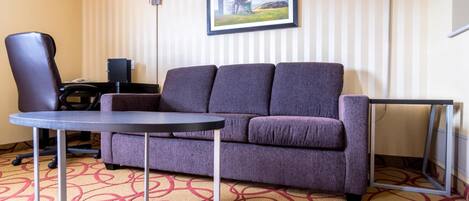 A room with a purple sofa, a small round table, a chair, a lamp, and a framed picture on the wall.