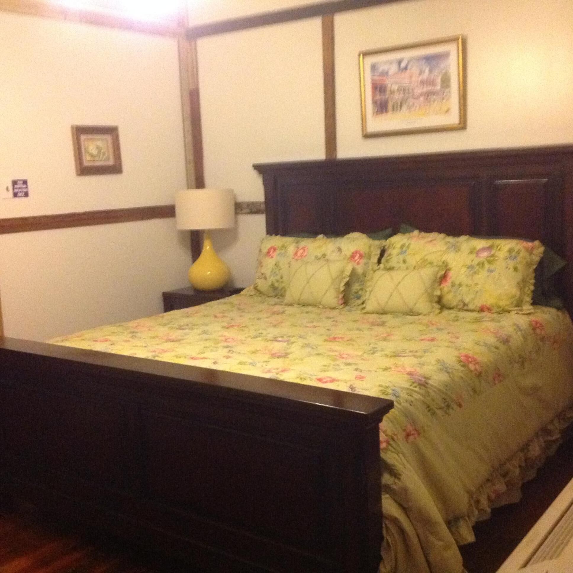 Un dormitorio con cabecera de cama de madera, ropa de cama con motivos florales, una mesita de noche con lámpara y un cuadro enmarcado en la pared.