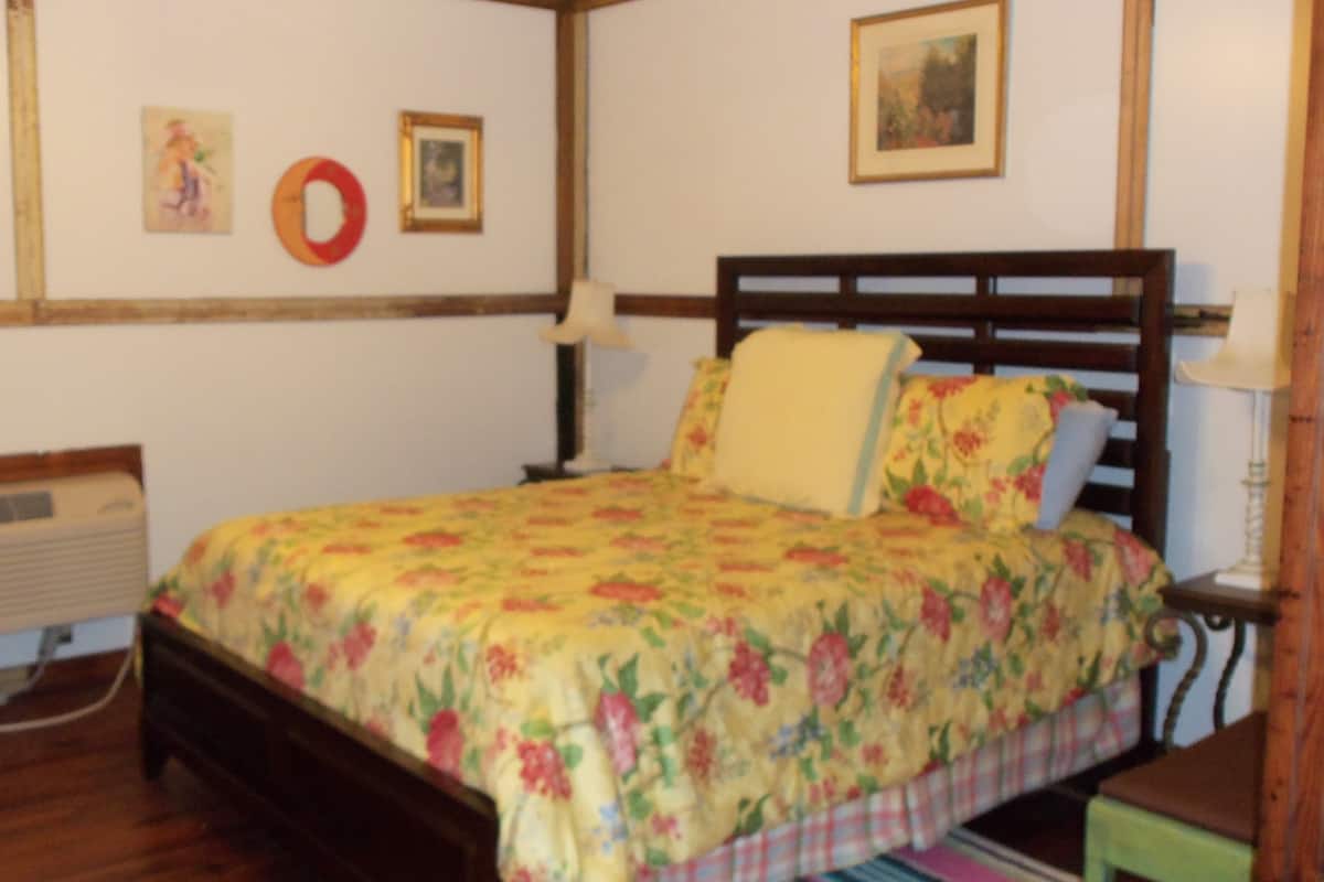 Ein Schlafzimmer mit einem hölzernen Bett, floralem Bettbezug, einem Nachttisch mit Lampe, einer Wandheizung und einem gerahmten Bild.