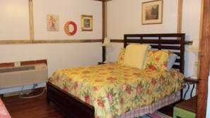 A bedroom with a wooden bed, floral bedding, a bedside table with a lamp, a wall-mounted heater, and a framed picture. - The Main Street Hotel (Yazoo City)