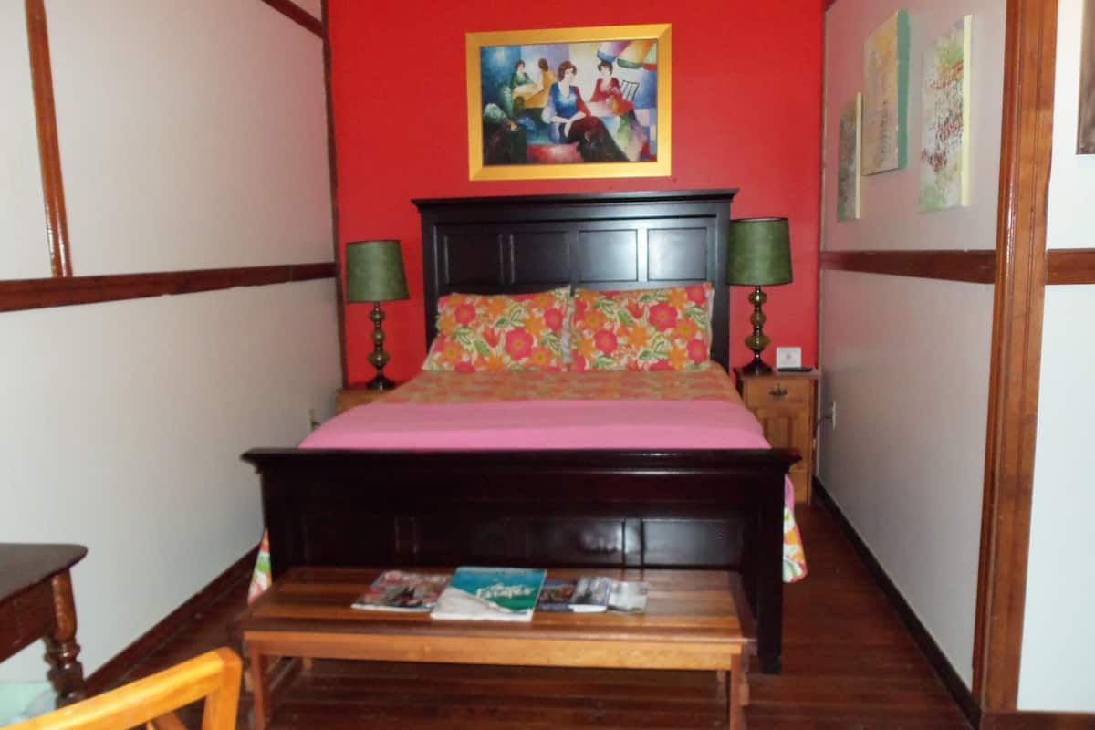 Ein Schlafzimmer mit einem hölzernen Bett, einer rosa Tagesdecke, blumigen Kissen und einem Bild an der Wand.