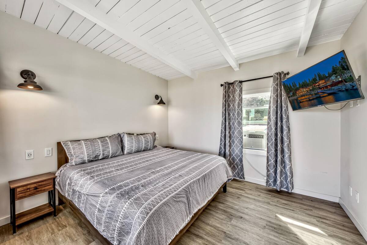 A bedroom with a bed, wooden nightstand, ceiling fan, wall-mounted TV, and window with curtains.