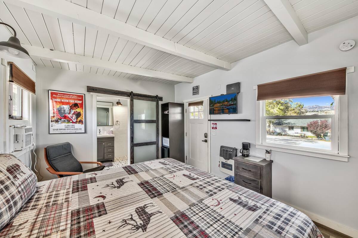 A bedroom with a plaid bedspread, a desk with a coffee maker, a window with a view, and a poster on the wall.