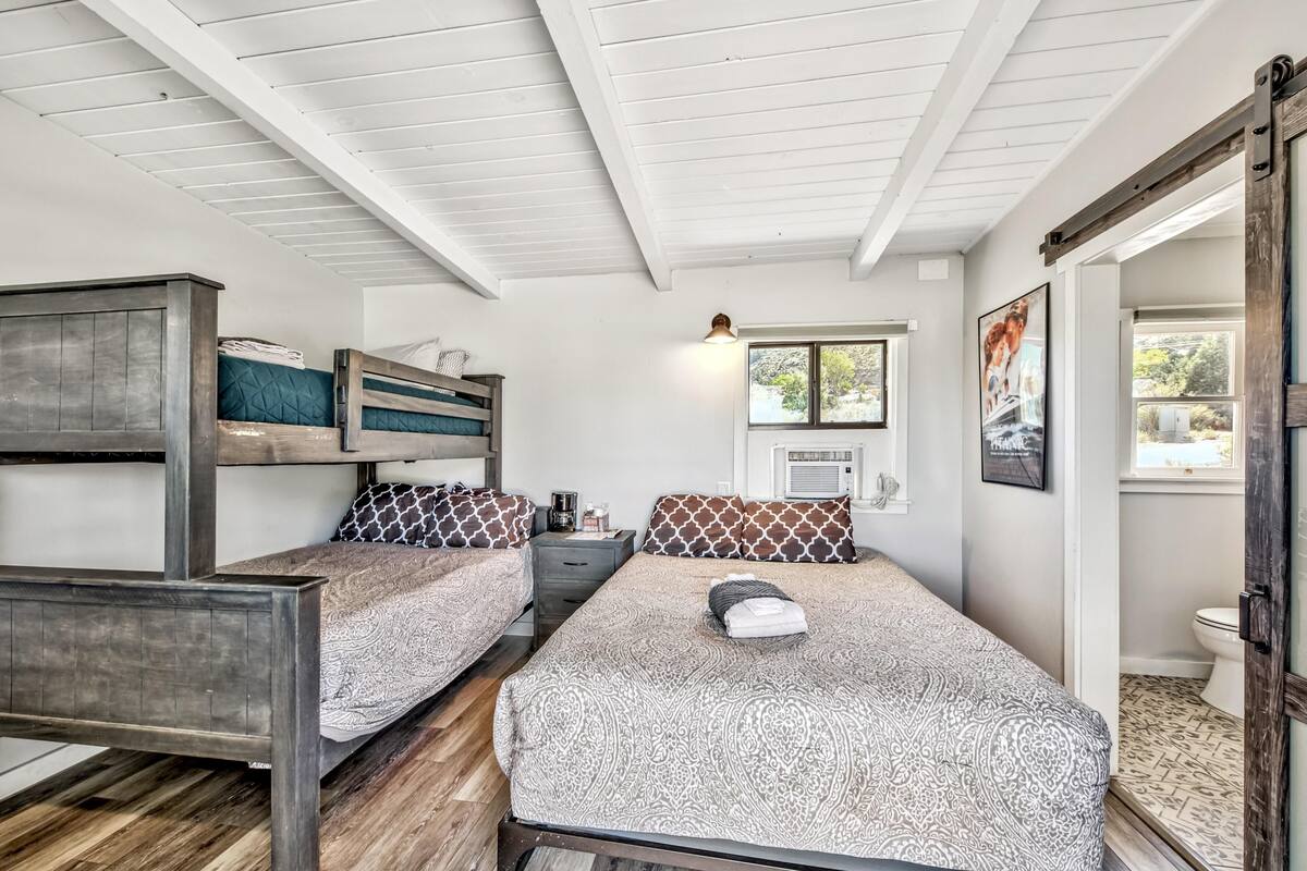 A room with bunk beds, a desk, a chair, a window with a view, and a door leading to a bathroom.