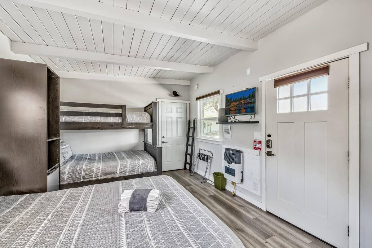 A room with two bunk beds, a desk, a chair, a window with blinds, a door, and a mounted TV.