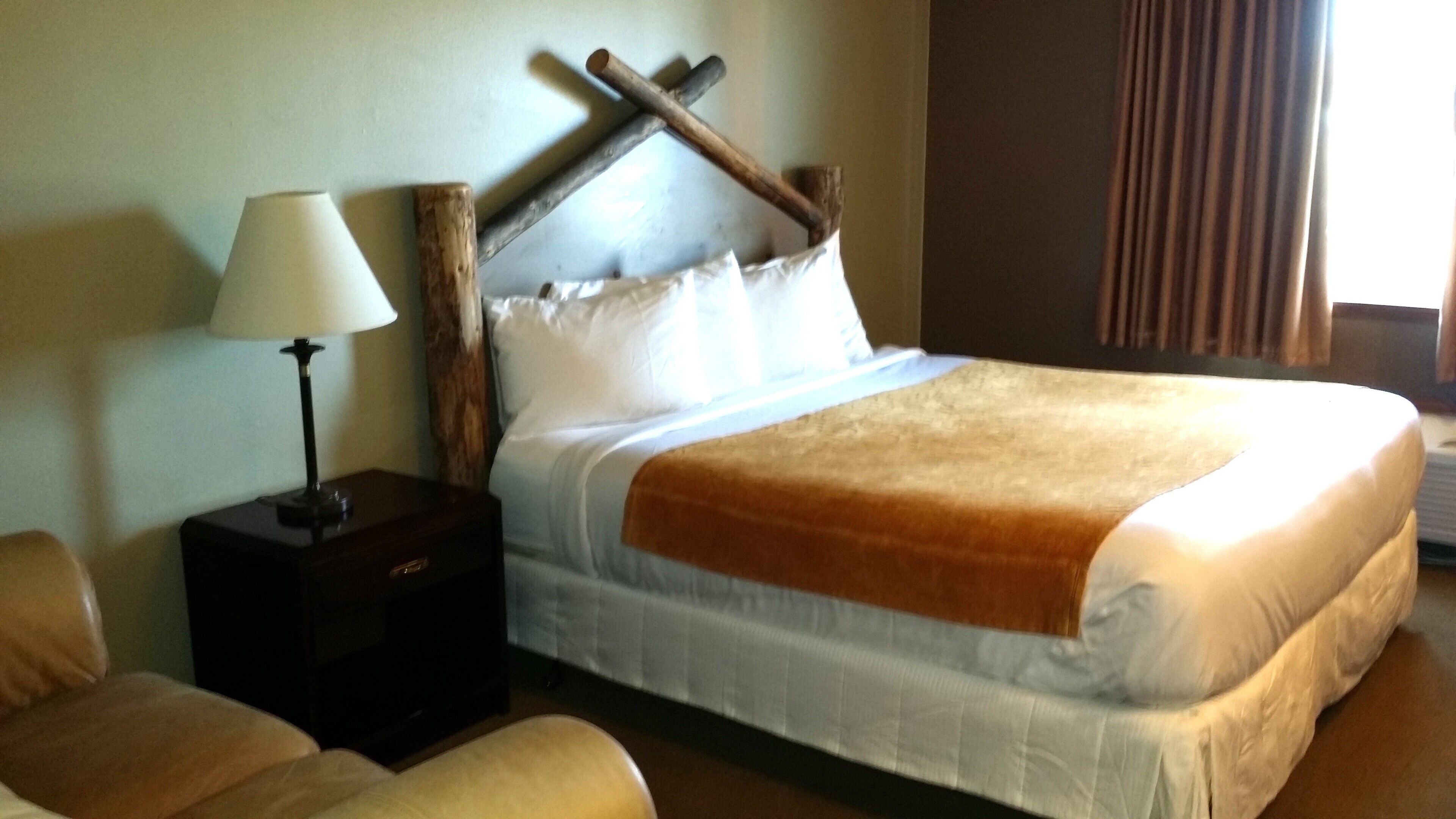 Ein Hotelzimmer mit Bett, Nachttisch mit Lampe, Holz-Kopfteil und einem Fenster mit Vorhängen.