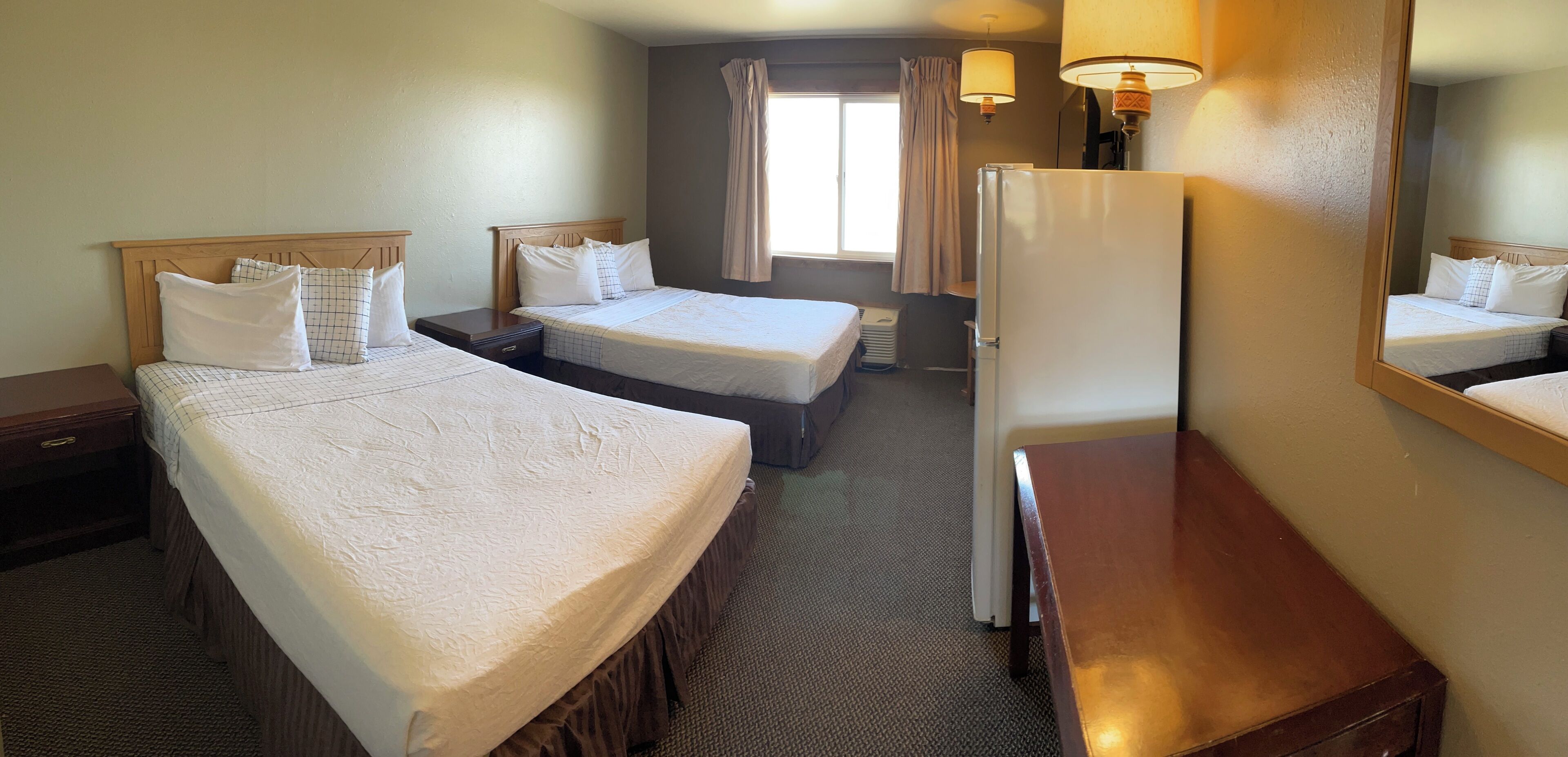 A hotel room with two single beds, a wooden nightstand, a desk, a refrigerator, and a window with curtains.
