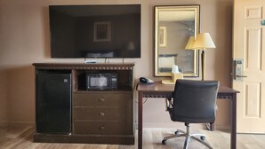 A hotel room with a flat-screen TV, a microwave, a desk with a chair, a lamp, and a door.