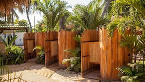 Una cerca de madera con un portón tejido, rodeada de plantas tropicales y un camino de madera.