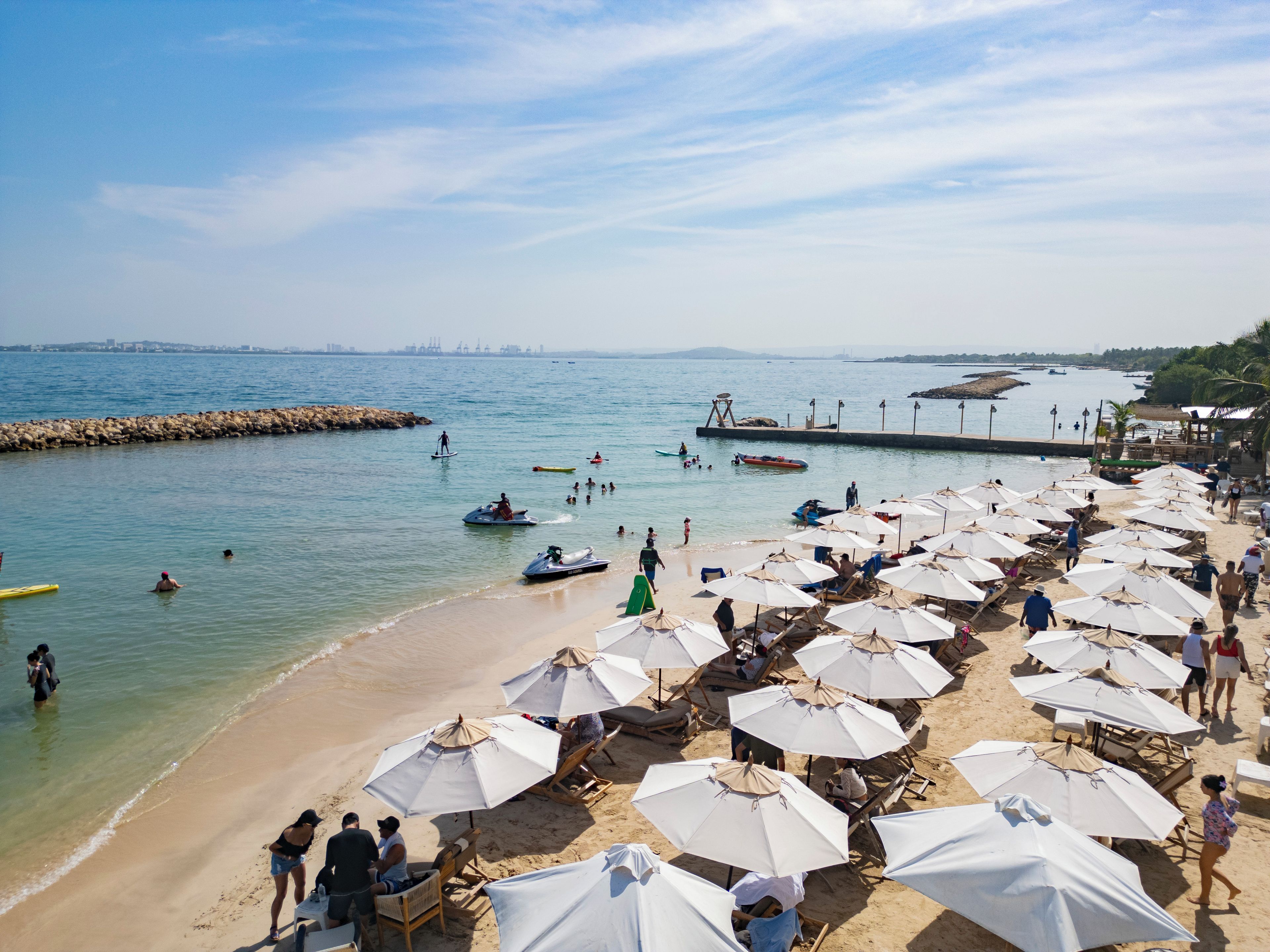 A beach with rows of umbrellas and sun loungers, a rocky breakwater, and people in the water.