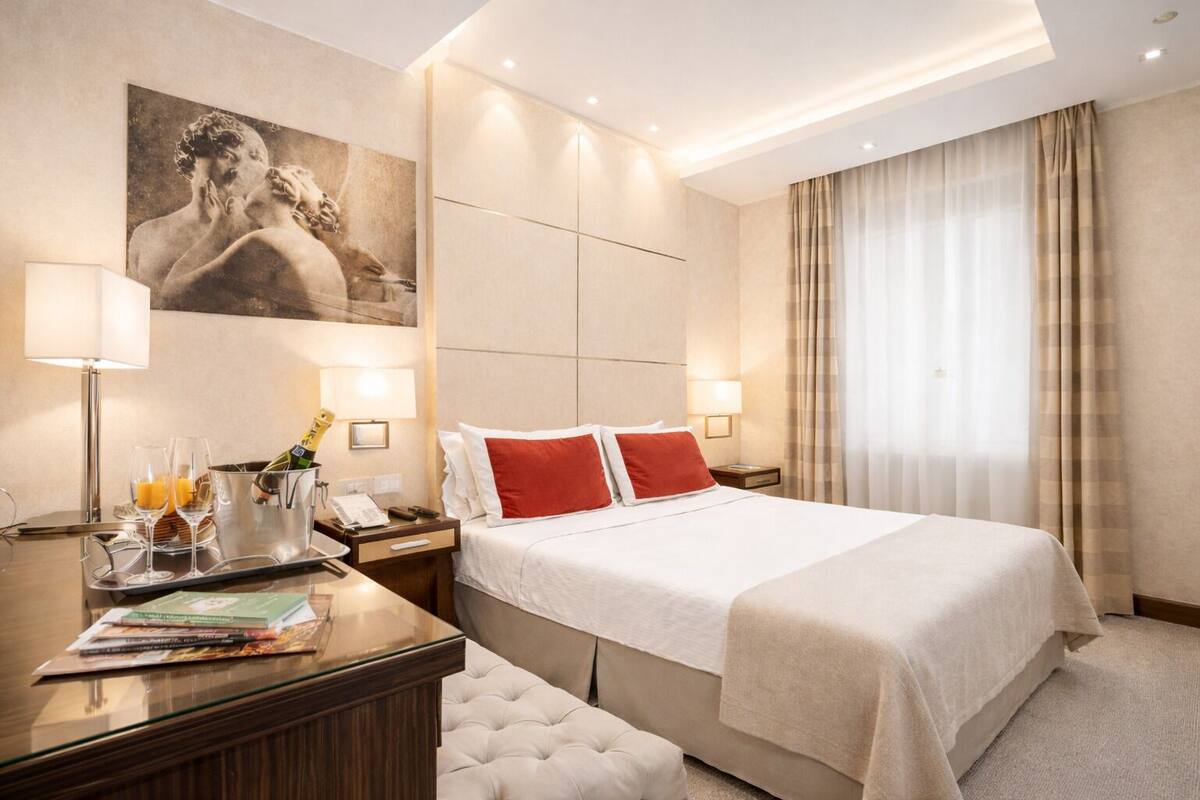 Una camera d'albergo con un letto grande, una scrivania con una lampada, un quadro appeso e una finestra con tende.