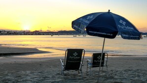 Two beach chairs under a blue umbrella with the Paradise logo, a sandy beach, and a sunset over the ocean.