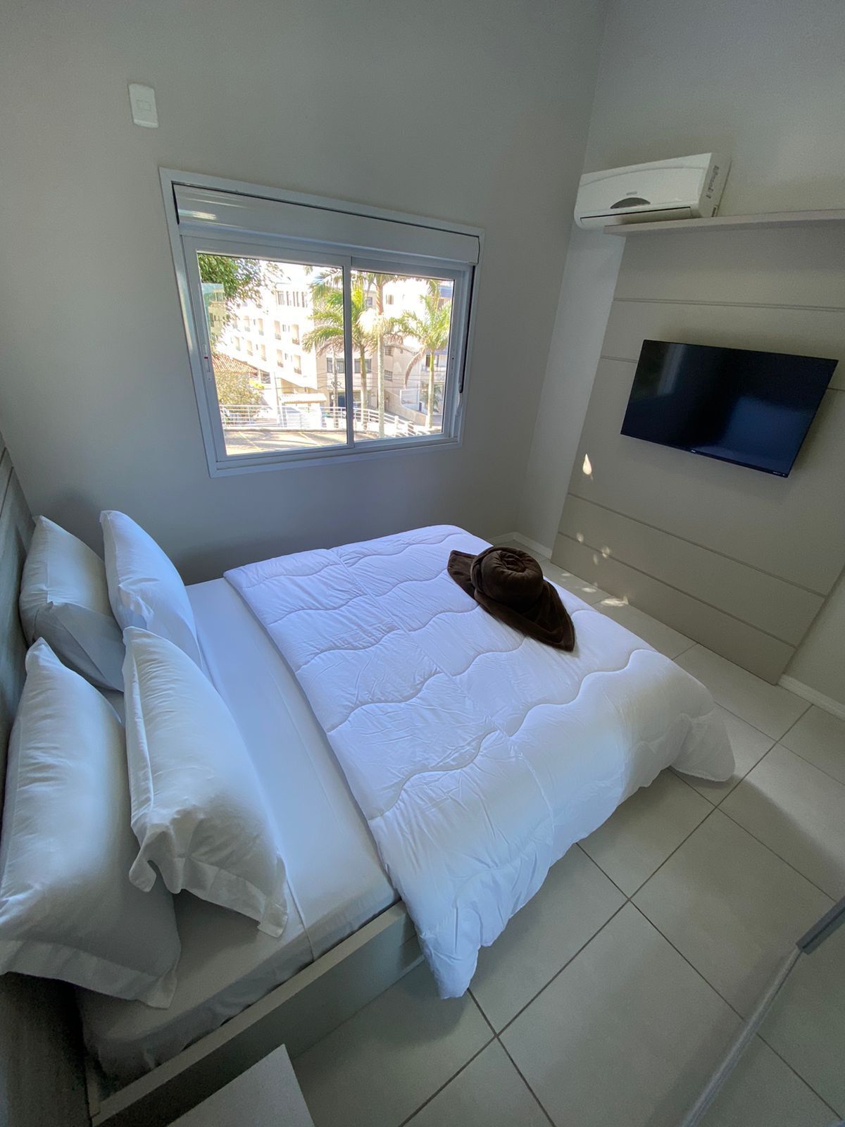Een slaapkamer met een bed, kussens, een raam en een wandtelevisie.