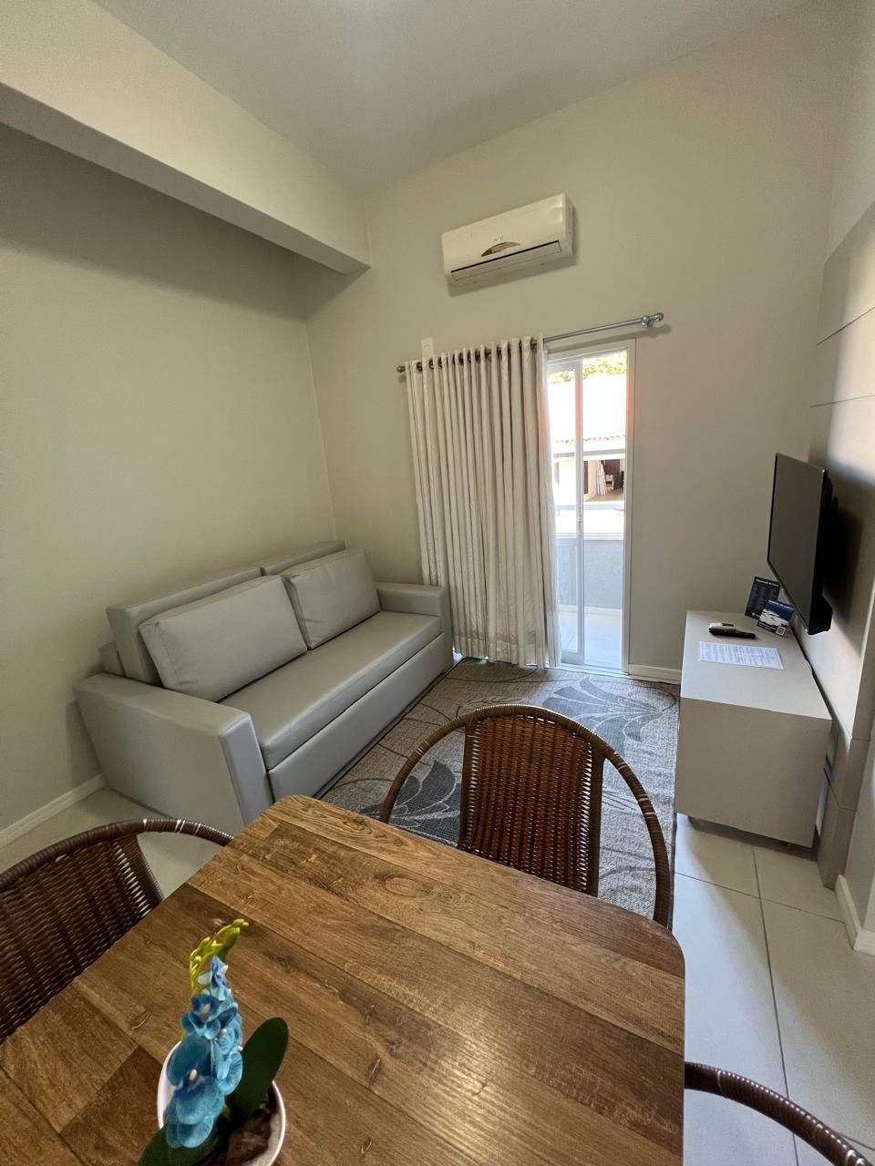 A living room with a sofa, a wooden dining table, wicker chairs, a TV, and an air conditioning unit.