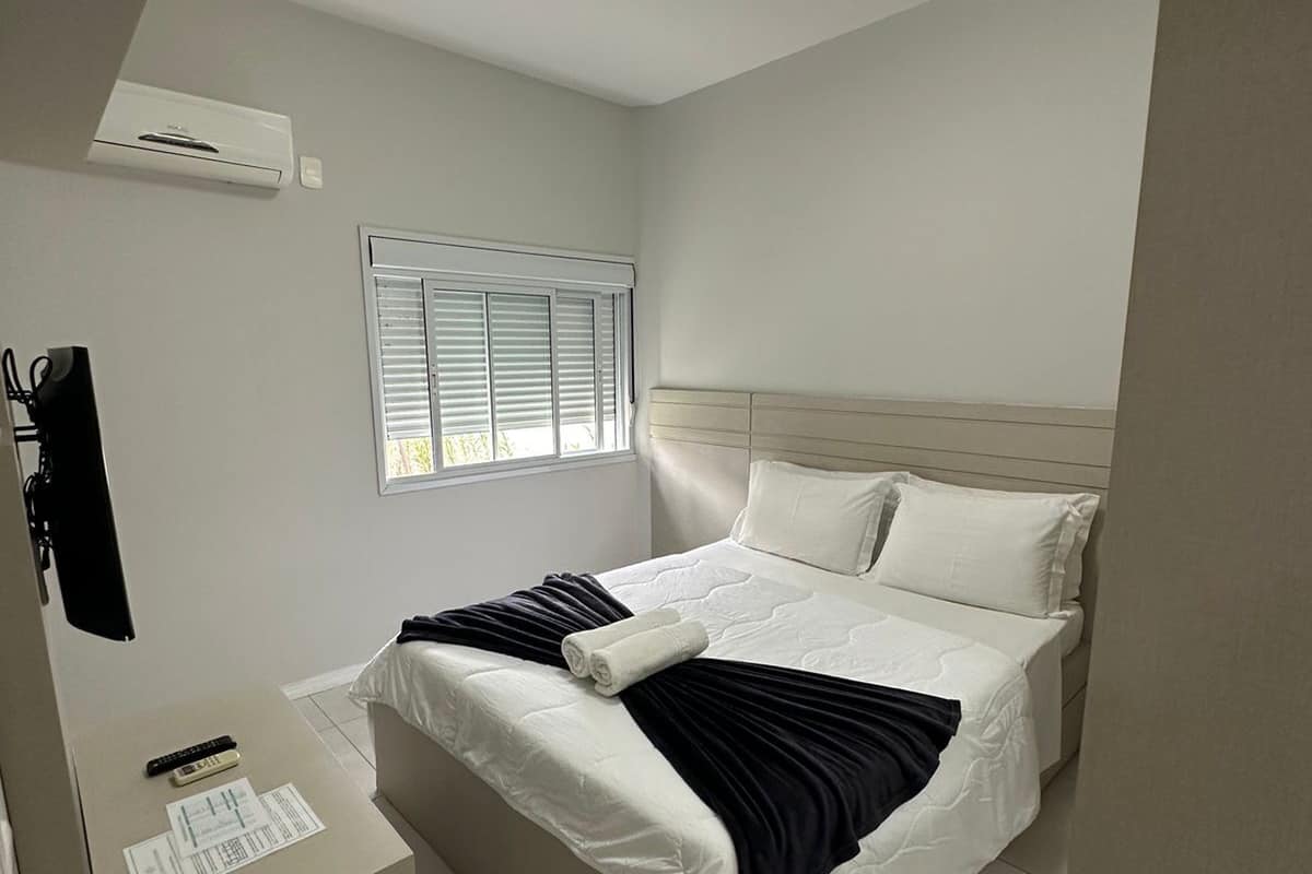 A bedroom with a bed, a bedside table, a window with blinds, and a ceiling light.