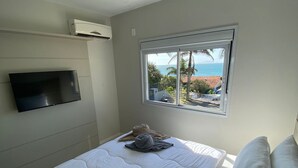 A bedroom with a bed, a TV mounted on the wall, an air conditioning unit, and a window with a view of the ocean and palm trees.