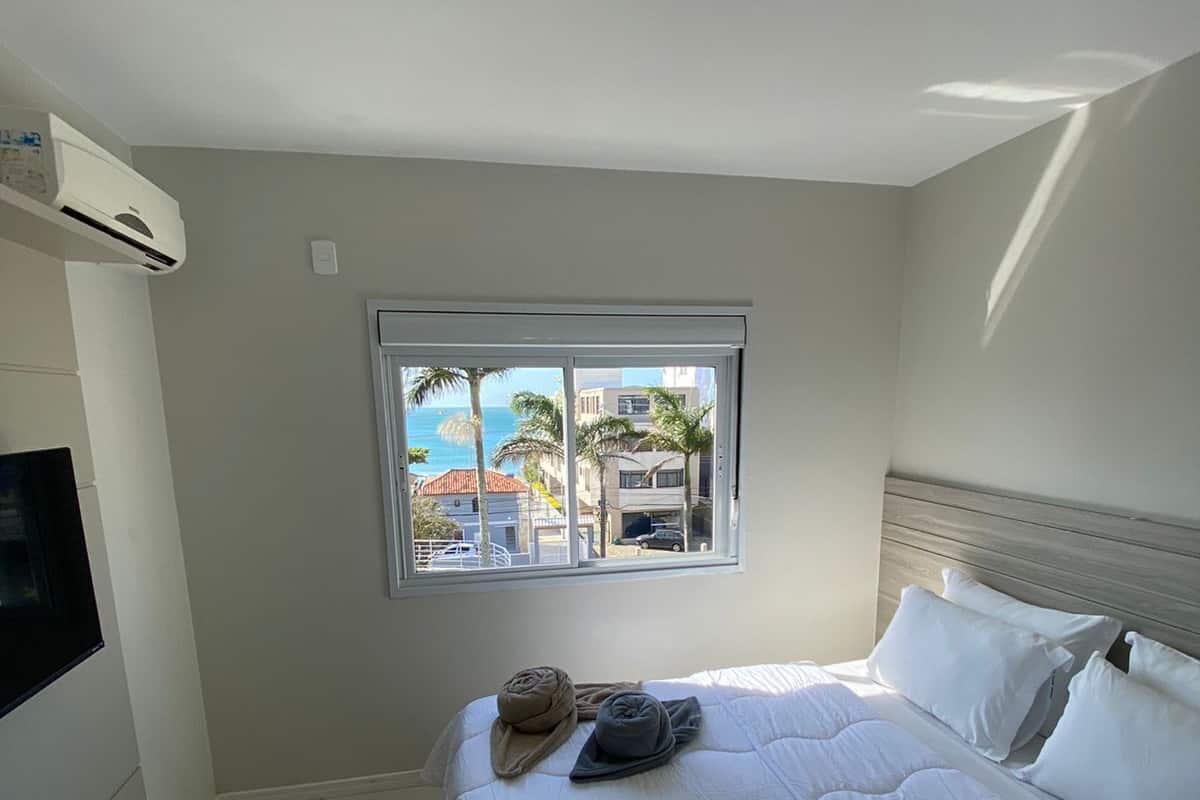 A hotel room with a bed, a TV, a window with a view of palm trees and buildings, and an air conditioning unit.