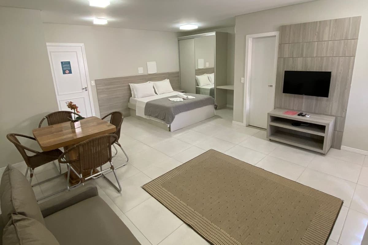 A modern hotel room with a bed, a TV, a dining area with a table and chairs, and a large window.