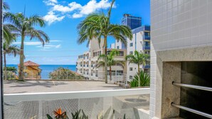 A hotel balcony with a view of the ocean and palm trees, a tiled floor, and a stone wall with a metal railing.