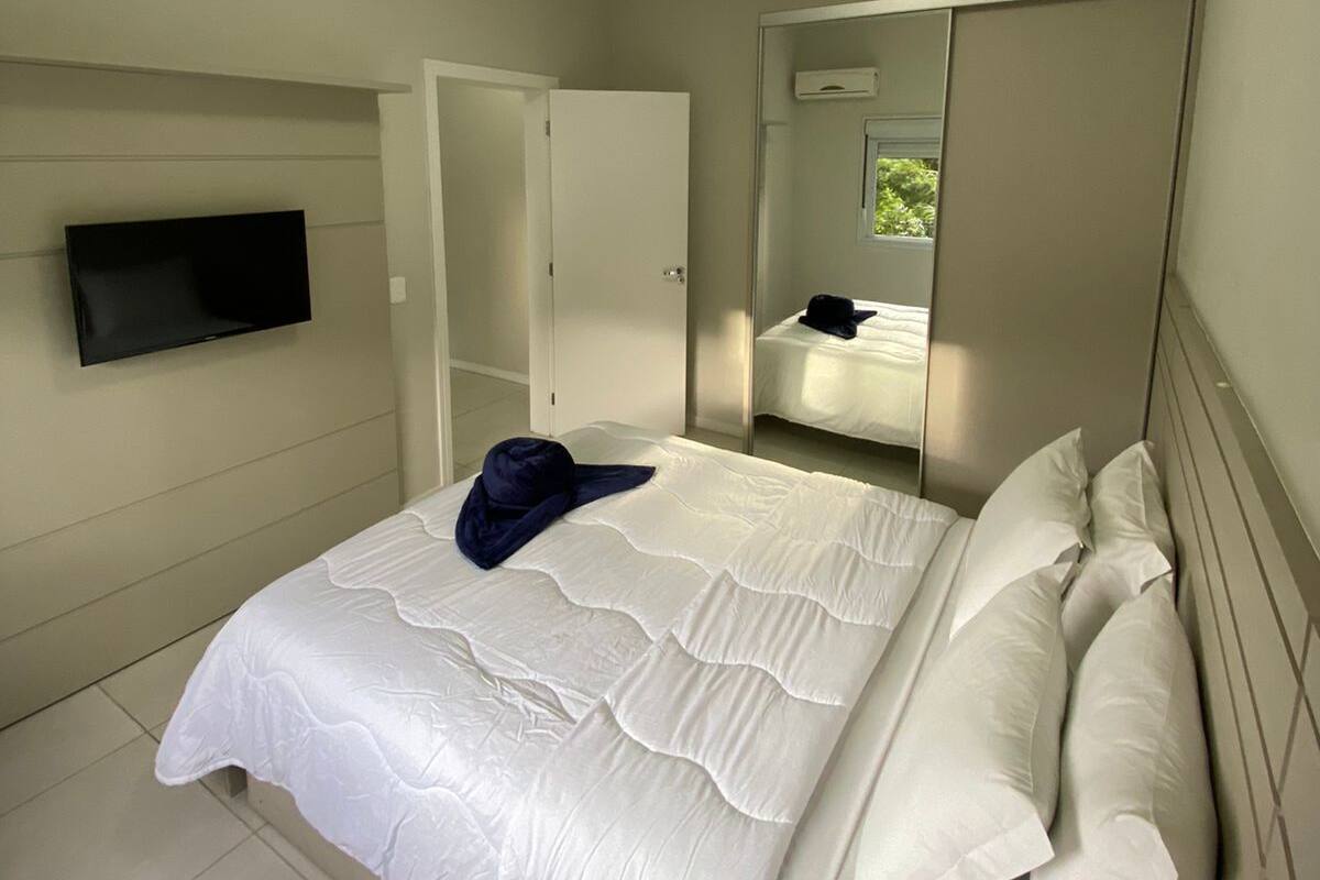 A neatly arranged bedroom with a bed, a flat-screen TV mounted on the wall, a wardrobe, and a mirror reflecting the room.