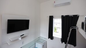Une chambre d’hôtel équipée d’un téléviseur à écran plat fixé au mur, d’une petite table en verre, d’une fenêtre avec des rideaux noirs et d’un appareil de climatisation.