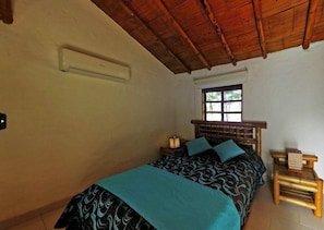 A bedroom with a wooden ceiling, a bed with patterned bedding, a nightstand with a lamp, and a window with a view of greenery.