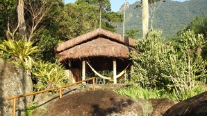 Une cabane au toit de chaume, dotée d’une balustrade en bois, entourée de gros rochers et d’une végétation luxuriante.