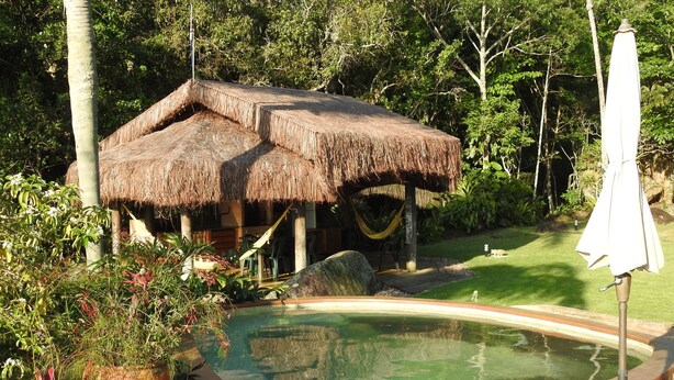 Une construction à toit de chaume, dotée d’une piscine et d’une terrasse en bois, entourée d’une végétation luxuriante.