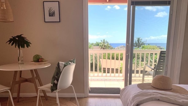 A bedroom with a bed, a chair, a small table with a plant, and a view of the ocean through a sliding door. - Maui Garden Oasis (Lahaina)
