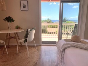 A bedroom with a bed, a chair, a small table with a plant, and a view of the ocean through a sliding door. - Maui Garden Oasis (Lahaina)