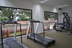 Een sportschool met loopbanden, een hometrainer en een krachttrainingapparaat. Grote ramen bieden uitzicht op een straat en bomen.