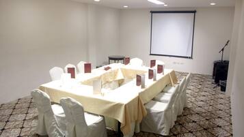 Un salón de conferencias con una larga mesa, sillas blancas, una pantalla de proyector y un soporte para micrófono.