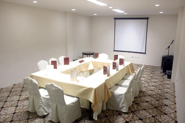 Un salón de conferencias con una larga mesa, sillas blancas, una pantalla de proyector y un soporte para micrófono.