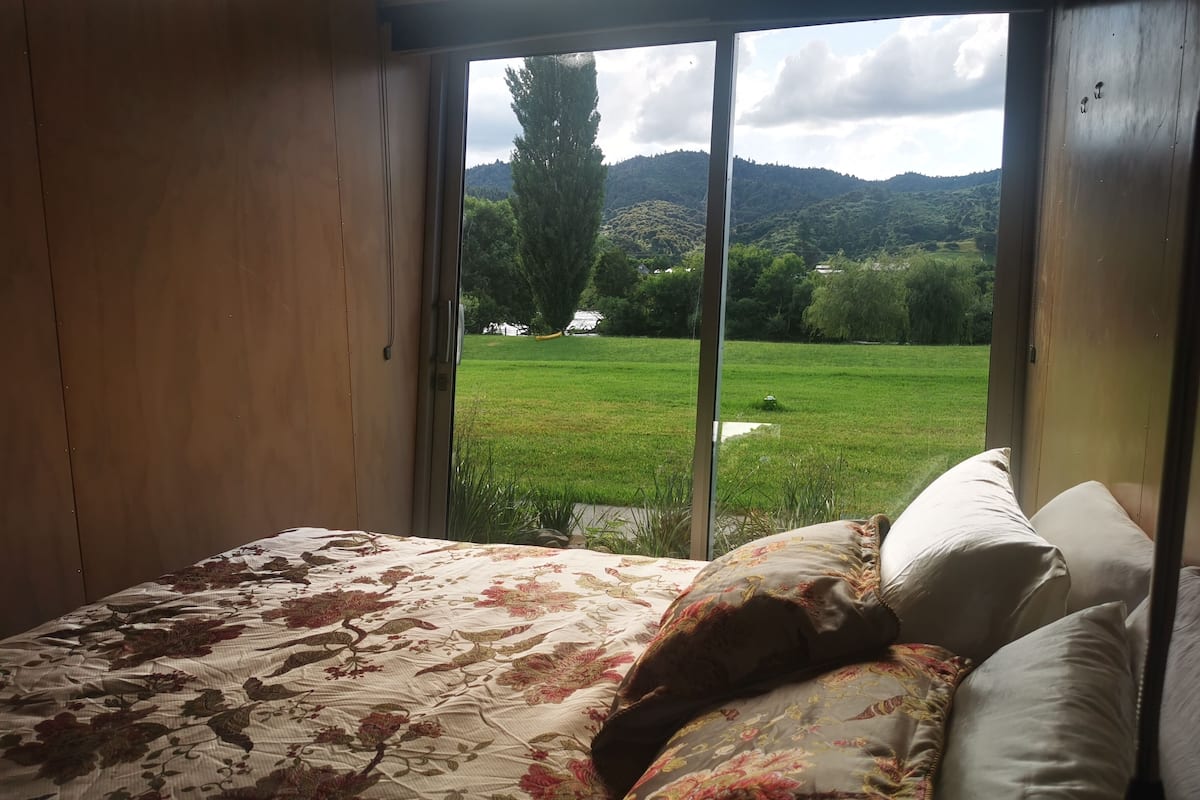 A bedroom with a bed, pillows, and a view of a green field and hills through a window.