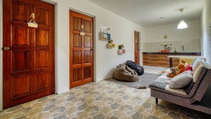 Sala de estar com um sofá, uma bolsa e um urso de pelúcia. Duas portas de madeira e uma cozinha ao fundo.