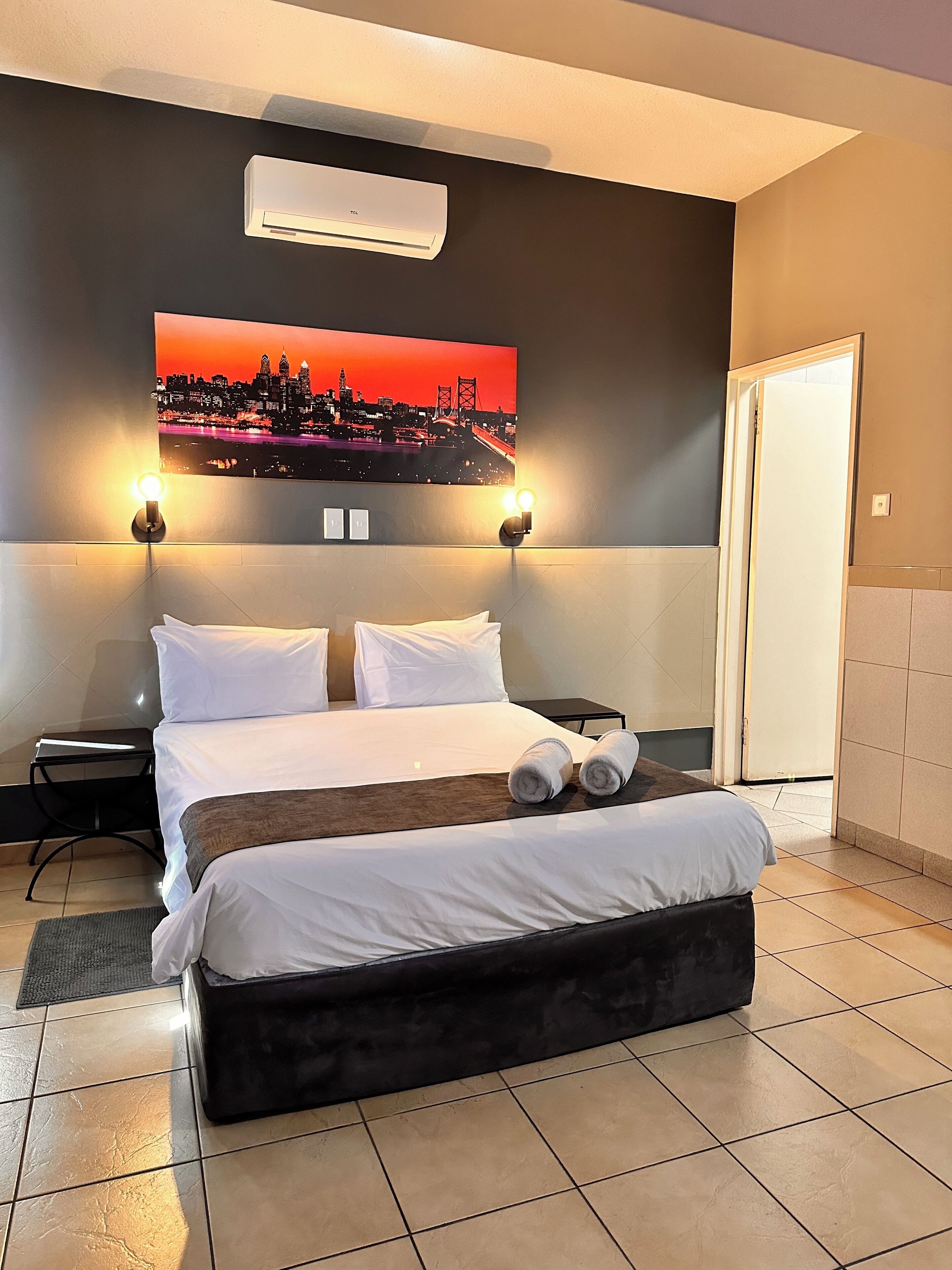 A hotel room with a large bed, two wall-mounted lights, a cityscape painting, and an air conditioning unit.