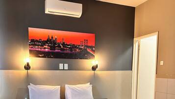 A hotel room with a large bed, two wall-mounted lights, a cityscape painting, and an air conditioning unit.