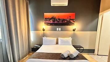 Quarto de hotel com uma cama grande, duas luminárias de cabeceira, um quadro com uma vista urbana na parede e uma porta à direita.