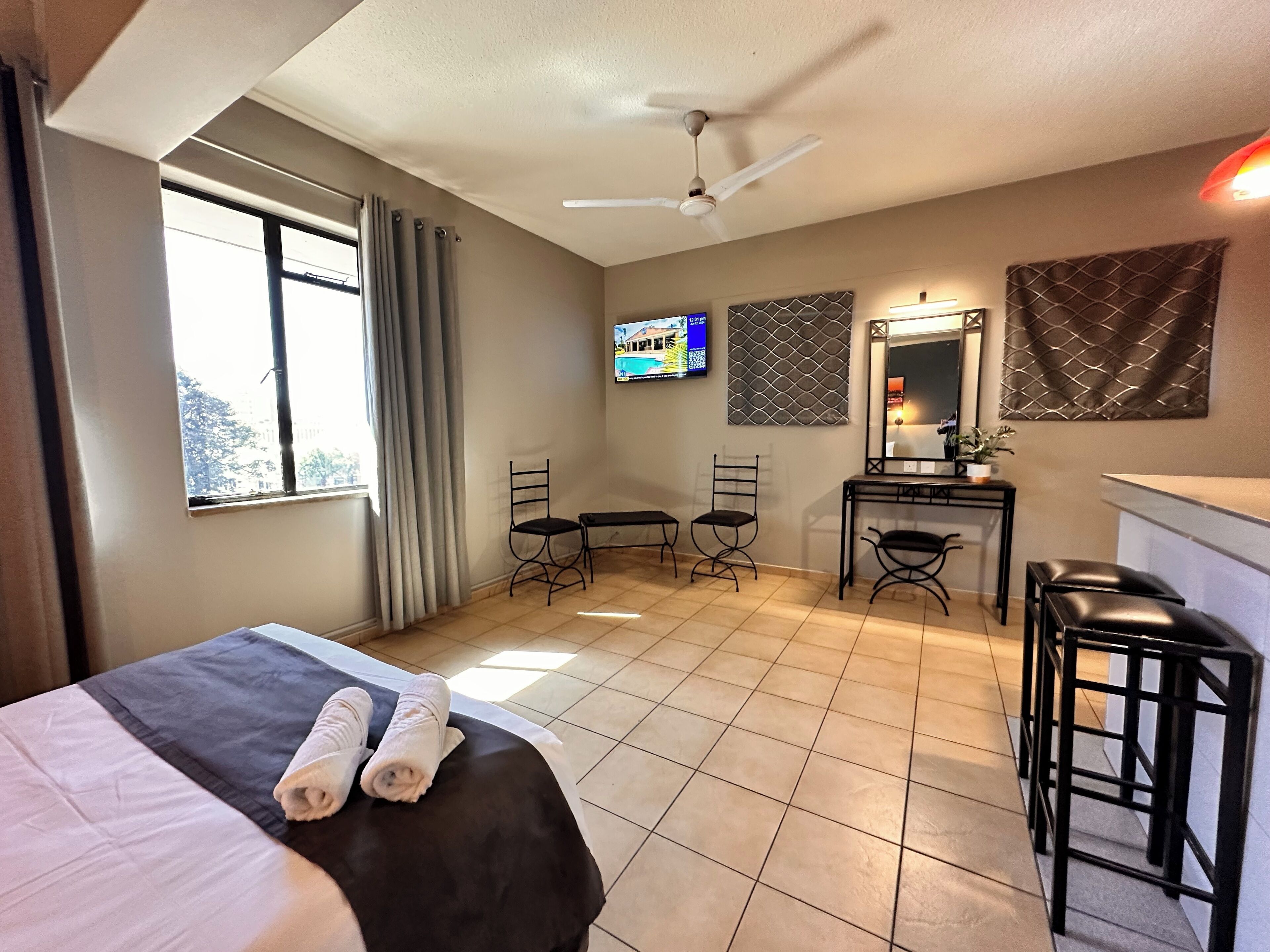 A hotel room with a bed, two chairs, a desk, a television, and a ceiling fan.