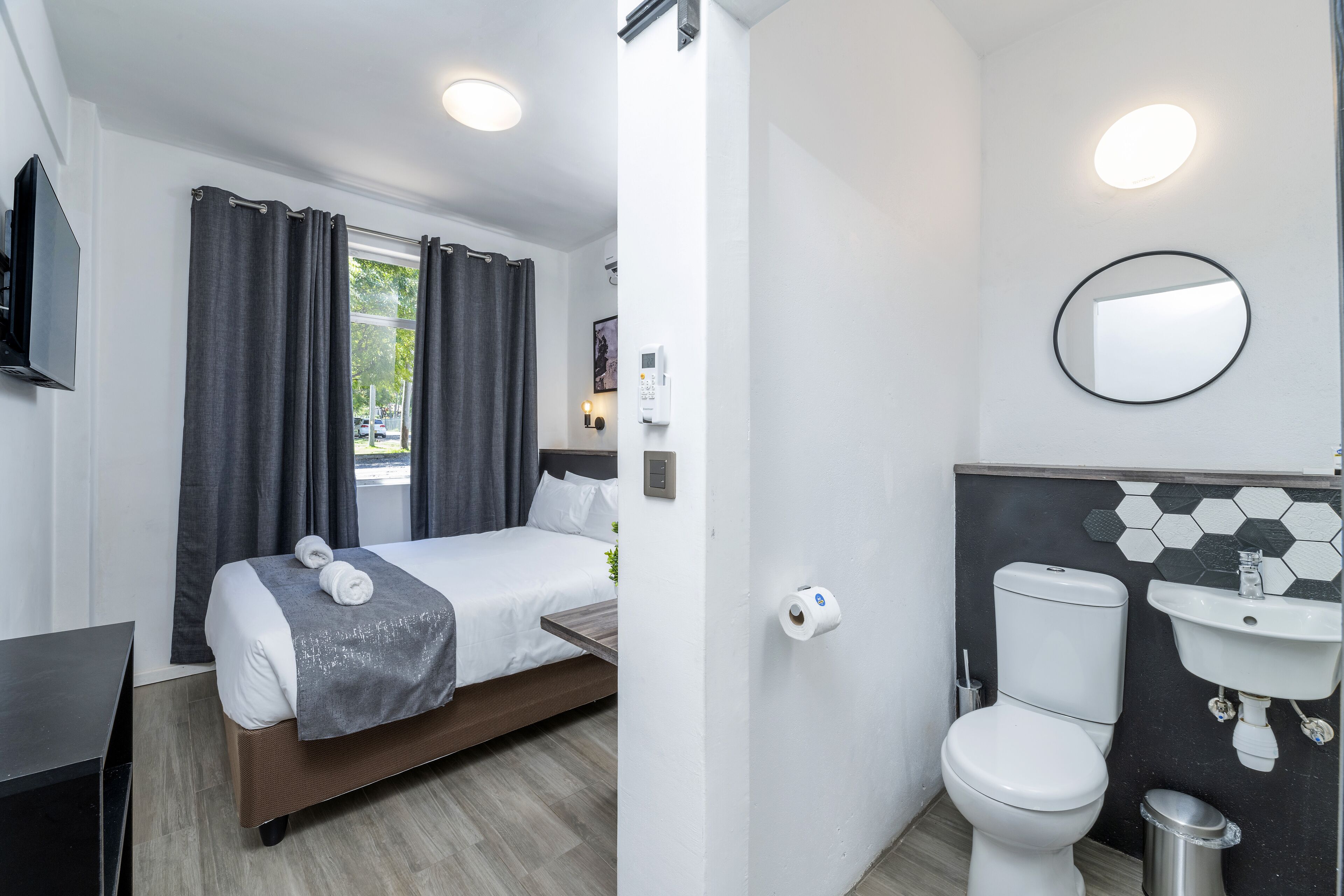 Ett modernt hotellrum med en säng, ett sängbord, en TV och ett badrum med toalett, handfat och spegel.