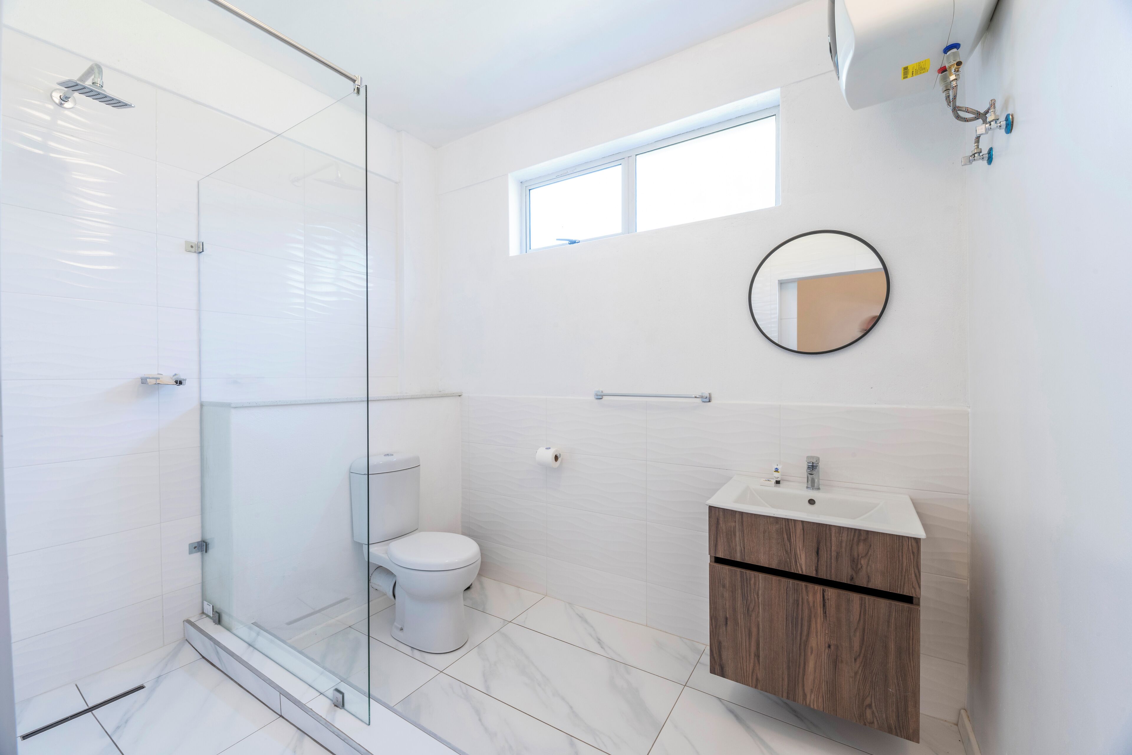 A modern bathroom with a glass shower enclosure, a toilet, a sink with a wooden cabinet, and a small window.
