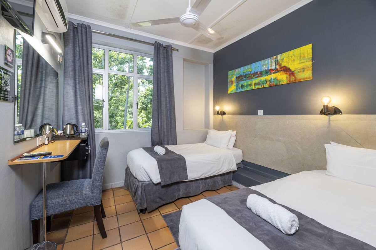 Habitación de hotel con dos camas, un escritorio con silla, una ventana con cortinas, un ventilador de techo y un cuadro en la pared.