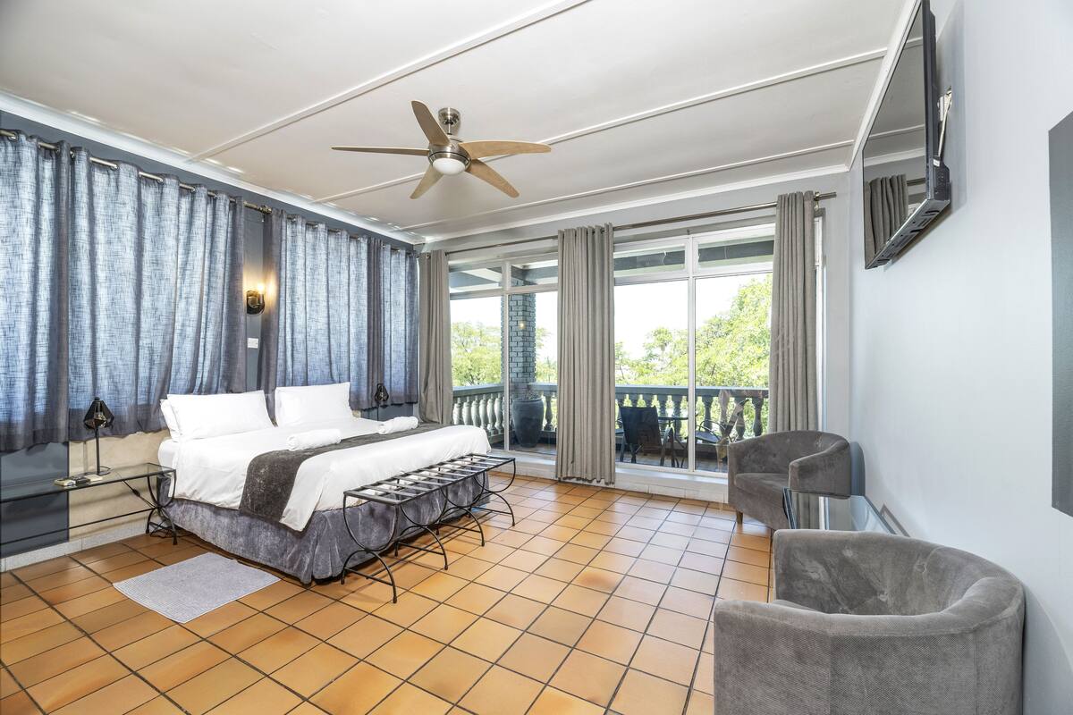 Un dormitorio espacioso con una cama grande, ventilador de techo, televisor montado en la pared y un balcón con vistas a zonas verdes.