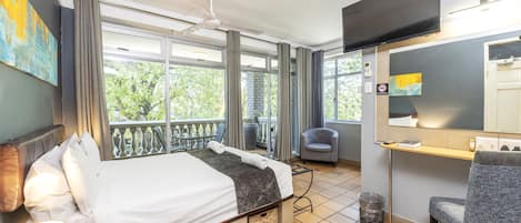 A hotel room with a bed, a chair, a desk, a television, and a balcony with a view of trees.