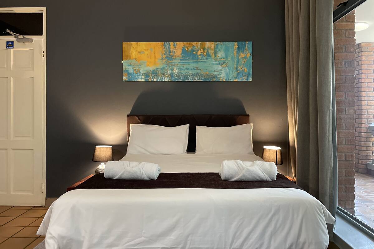 Una cama bien hecha con sábanas blancas, dos lámparas de noche y un cuadro abstracto grande en la pared.