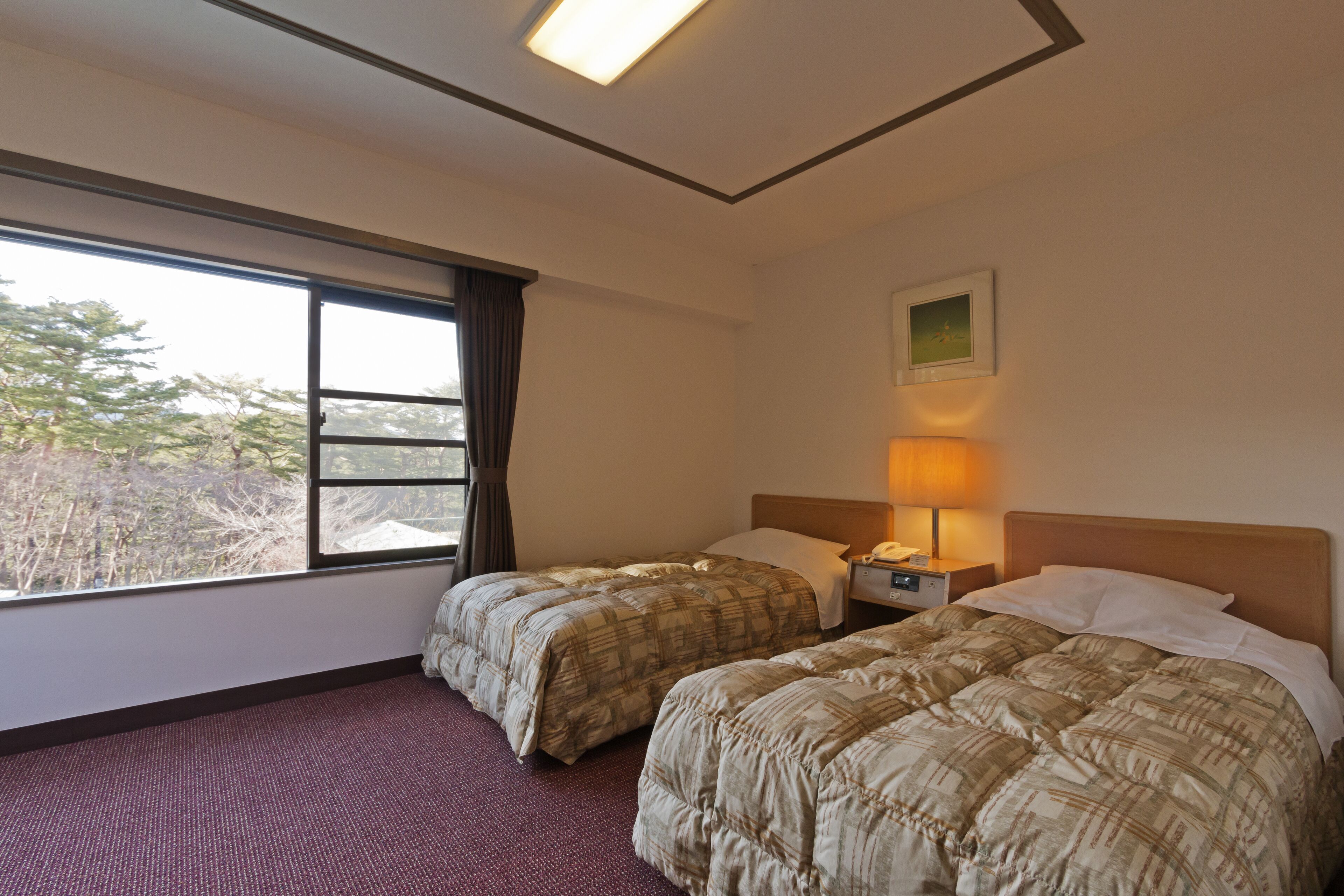 A hotel room with two beds, a window with curtains, a nightstand with a lamp, and a framed picture on the wall.