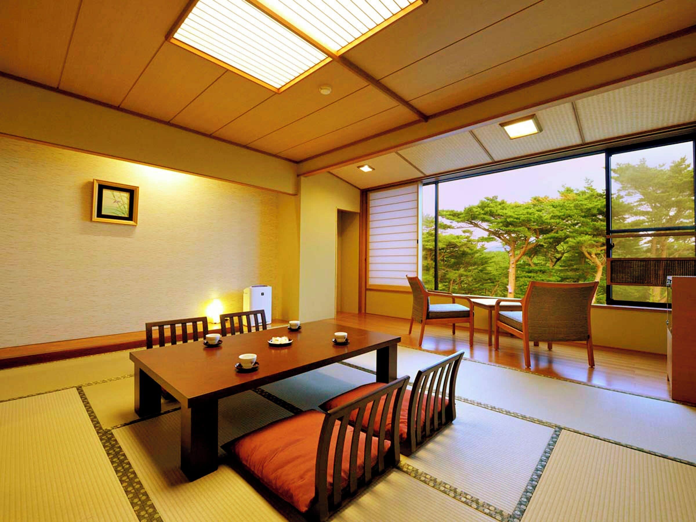 A traditional Japanese-style room with tatami flooring, a low table with cushions, and a view of trees through large windows.